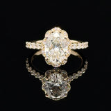 A yellow gold ring featuring a large oval-cut diamond in a decorative halo setting, with smaller diamonds along the band, displayed on a reflective black surface.