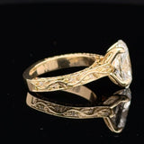 A yellow gold ring with an ornate engraved band and a large clear rectangular gemstone, shown on a reflective black surface.