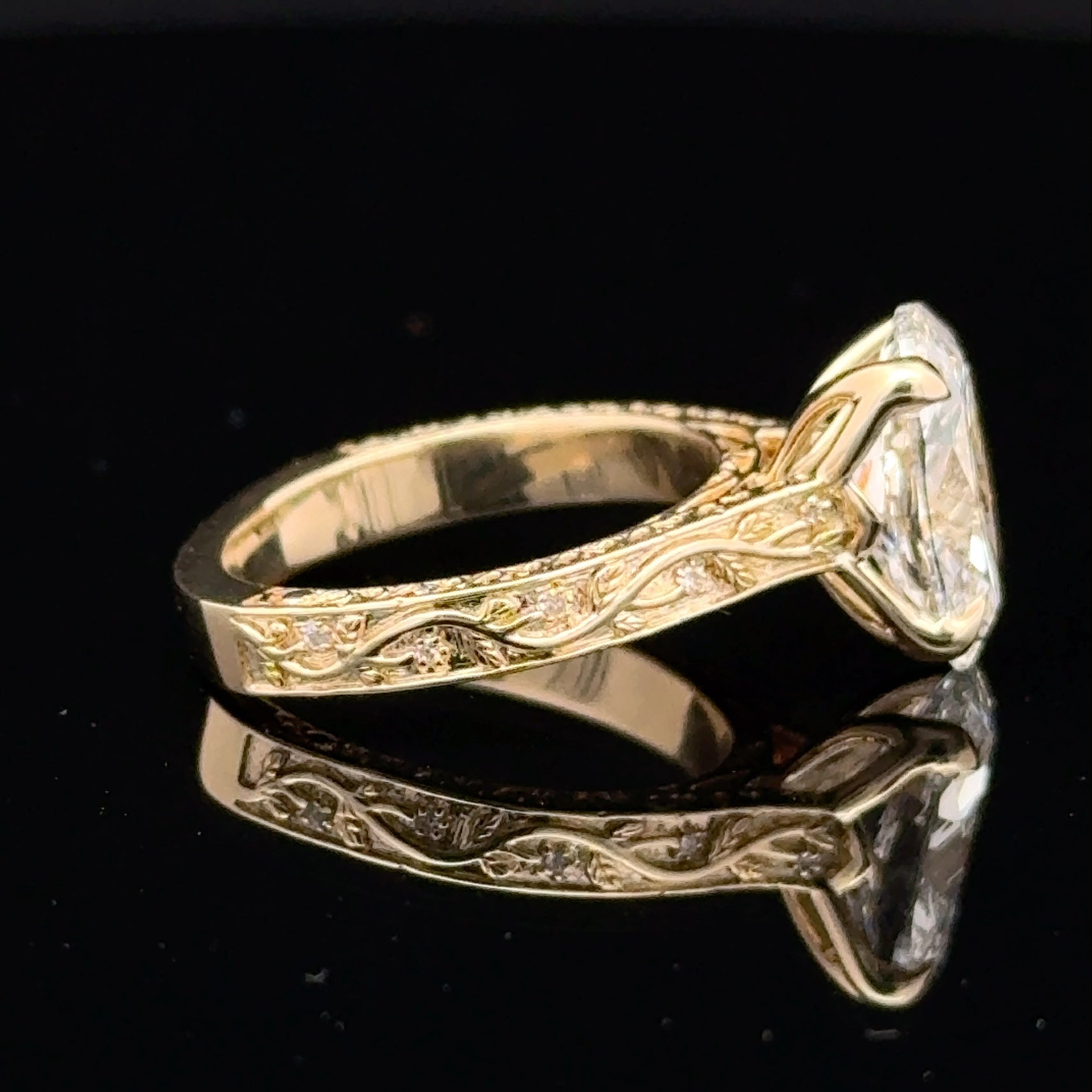 A yellow gold ring with an ornate engraved band and a large clear rectangular gemstone, shown on a reflective black surface.