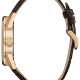 A side profile view of a watch with a rose gold case and a dark brown leather strap on a solid white background.