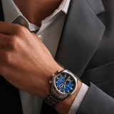Man wearing a silver watch with a blue face on his wrist, dressed in a gray suit.