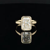 A yellow gold ring featuring a large rectangular emerald-cut diamond, displayed on a reflective black surface.
