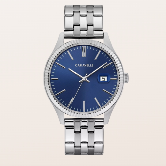 A luxury Caravelle wristwatch with a polished silver-tone stainless steel case and link bracelet, featuring a deep blue dial with gold-tone indices and a date window at the 3 o'clock position.