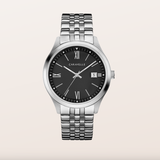 A Caravelle men's watch featuring a silver stainless steel case and link bracelet, a black dial with Roman numeral hour markers, and a date window at 3 o'clock, set against a solid light beige background.