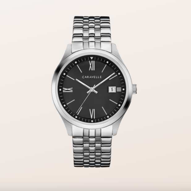 A Caravelle men's watch featuring a silver stainless steel case and link bracelet, a black dial with Roman numeral hour markers, and a date window at 3 o'clock, set against a solid light beige background.