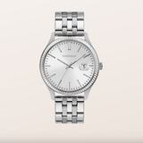 A silver-tone Caravelle wristwatch with a stainless steel link bracelet, a fluted bezel, and a white dial with a date window, centered on a light beige background.