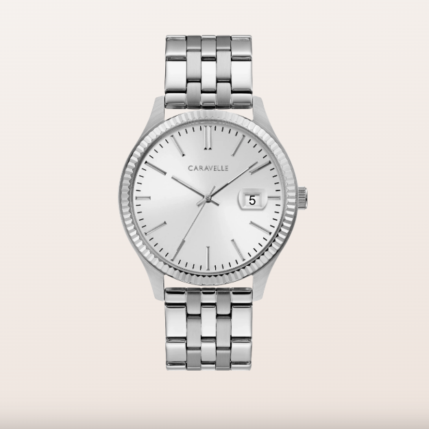 A silver-tone Caravelle wristwatch with a stainless steel link bracelet, a fluted bezel, and a white dial with a date window, centered on a light beige background.