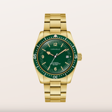 A Caravelle wristwatch with a gold-tone metal bracelet and case, featuring a dark green dial and bezel with gold-tone hour markers and a date window.