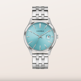 A Caravelle men's watch featuring a silver stainless steel bracelet and case, with a light turquoise blue dial and a date window at 3 o'clock, set against a solid light beige background.