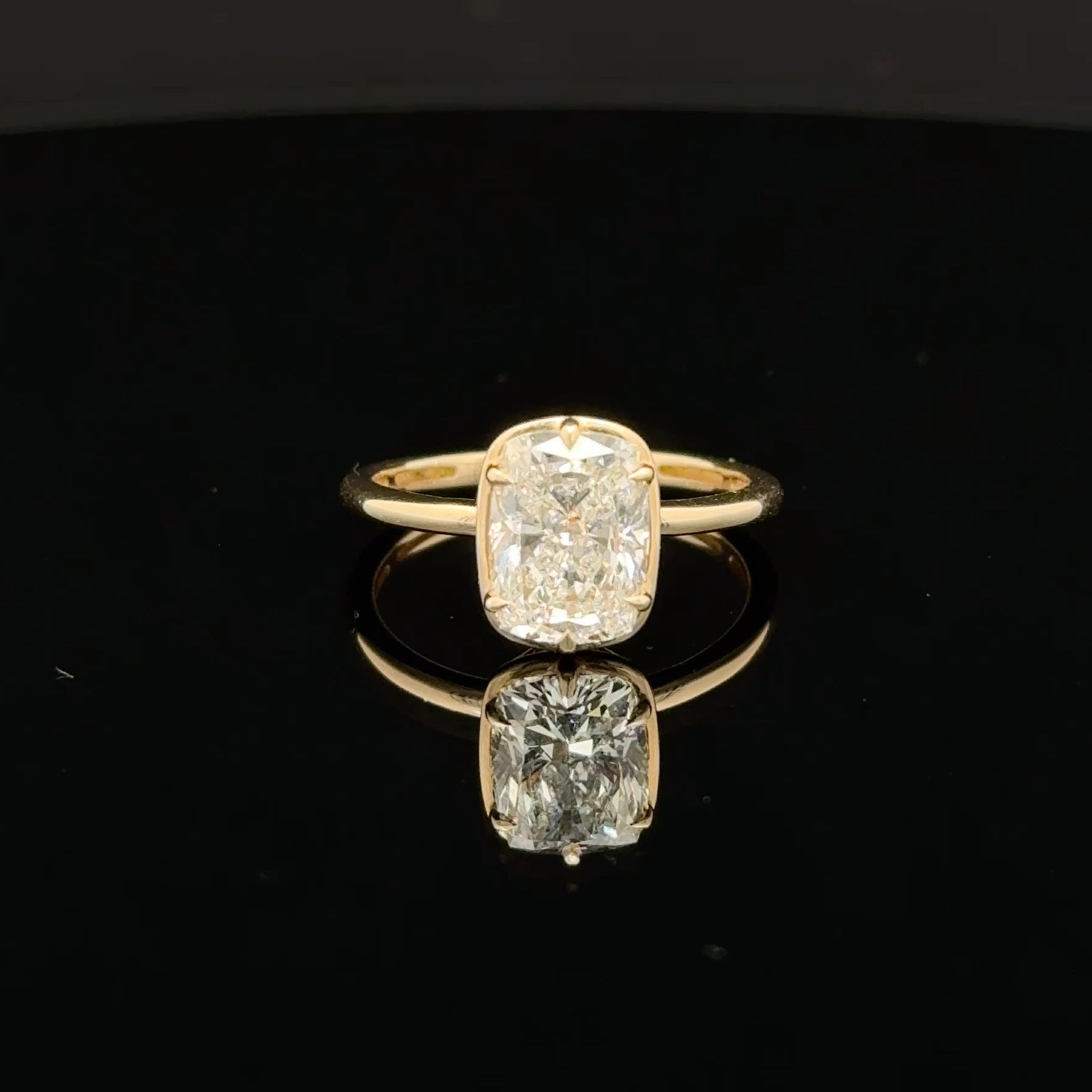 A yellow gold ring featuring a cushion-cut diamond, displayed on a reflective black surface.