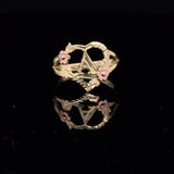 A gold ring featuring a central star motif, twisted vine details, and two small pink flower accents, displayed on a reflective black surface.