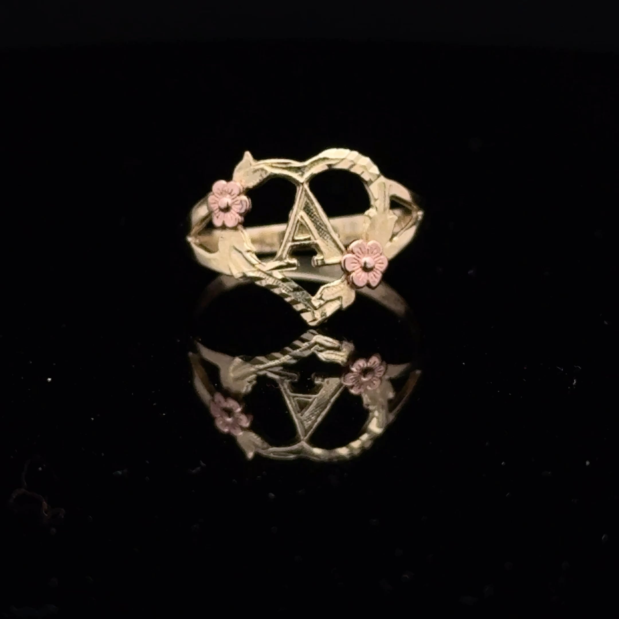A gold ring featuring a central star motif, twisted vine details, and two small pink flower accents, displayed on a reflective black surface.