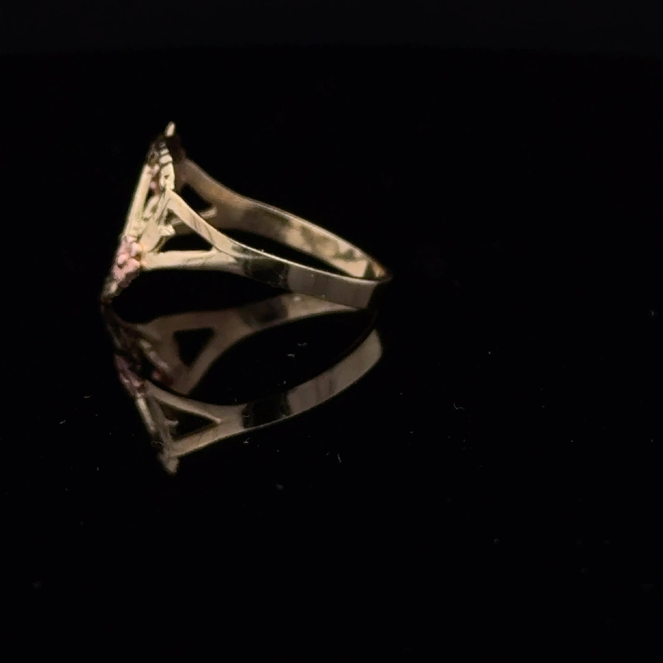 A gold ring with a triangular openwork design and small pink gemstone accents, displayed on a reflective black surface against a black background.