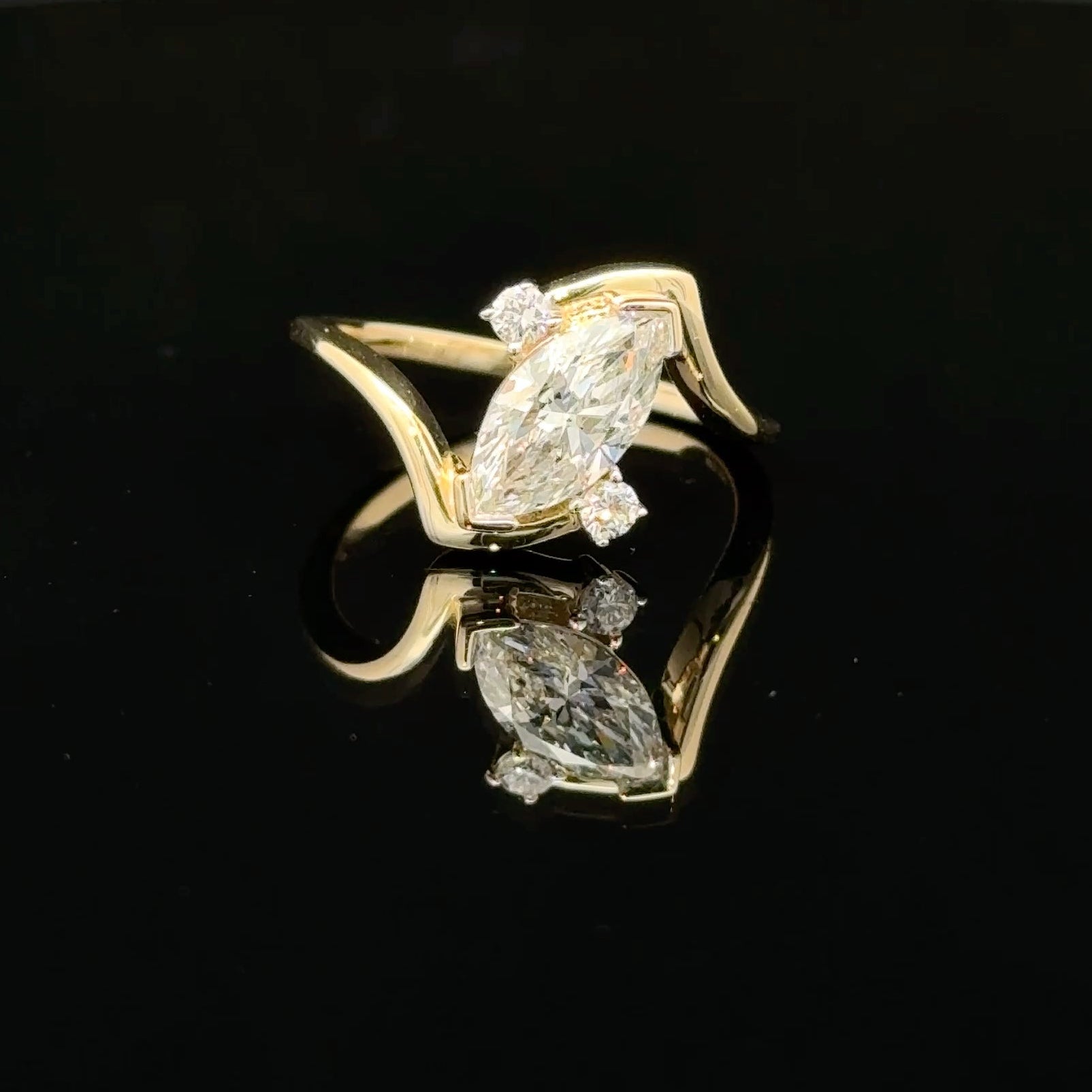 A yellow gold ring featuring a marquise-cut diamond center stone with smaller round diamonds on the shoulders, displayed on a reflective black surface.