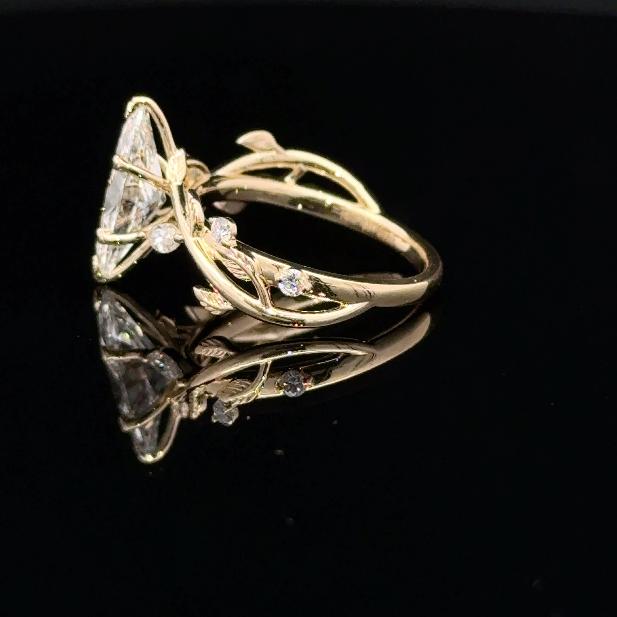 A gold ring with a large triangular clear gemstone and delicate leaf-like details on a black reflective background.