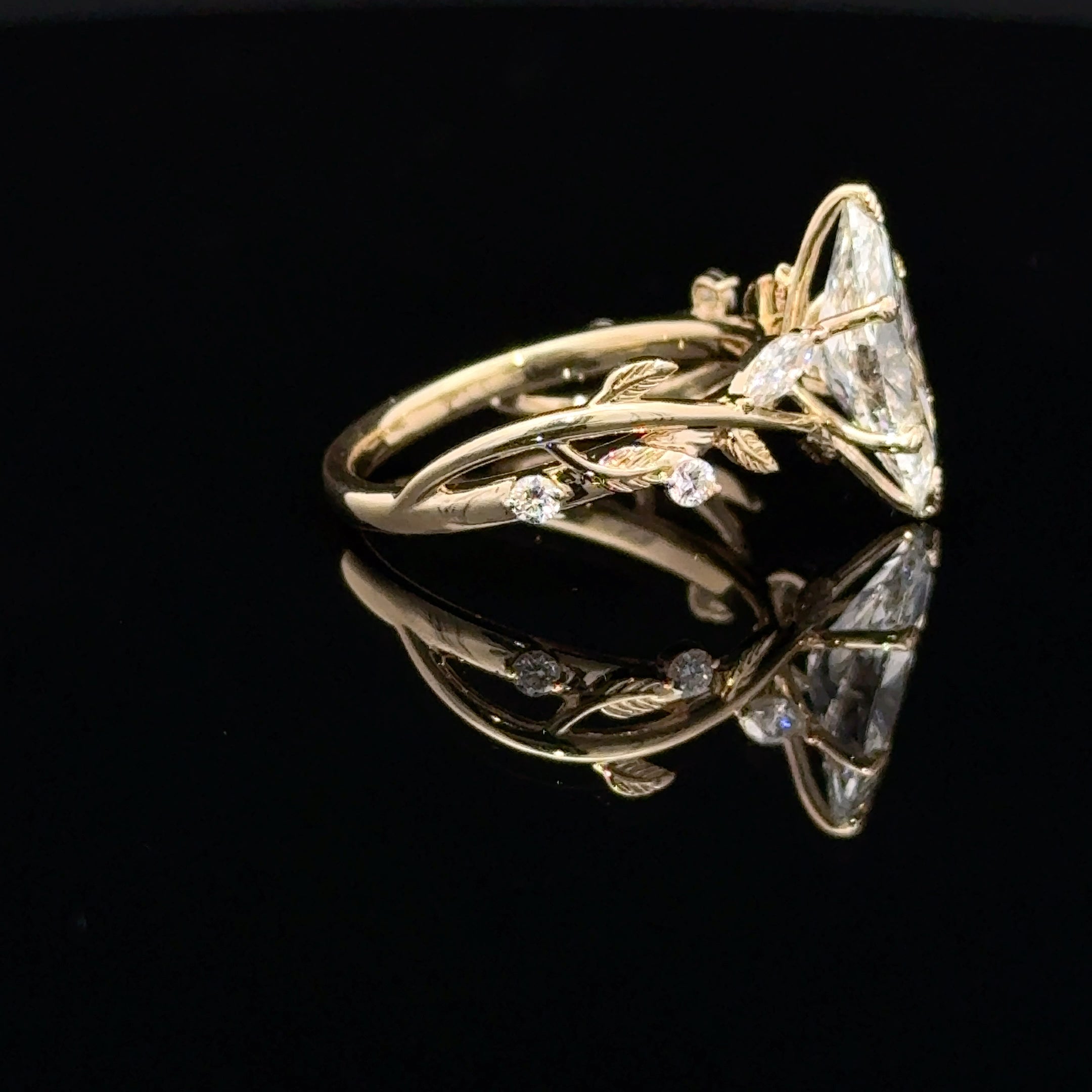 A gold ring featuring a large triangular clear gemstone and delicate leafy vine details, displayed on a reflective black surface.