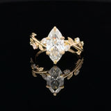 A gold ring featuring a large marquise-cut clear gemstone in the center, with smaller side stones and leaf-like details on a black reflective background.