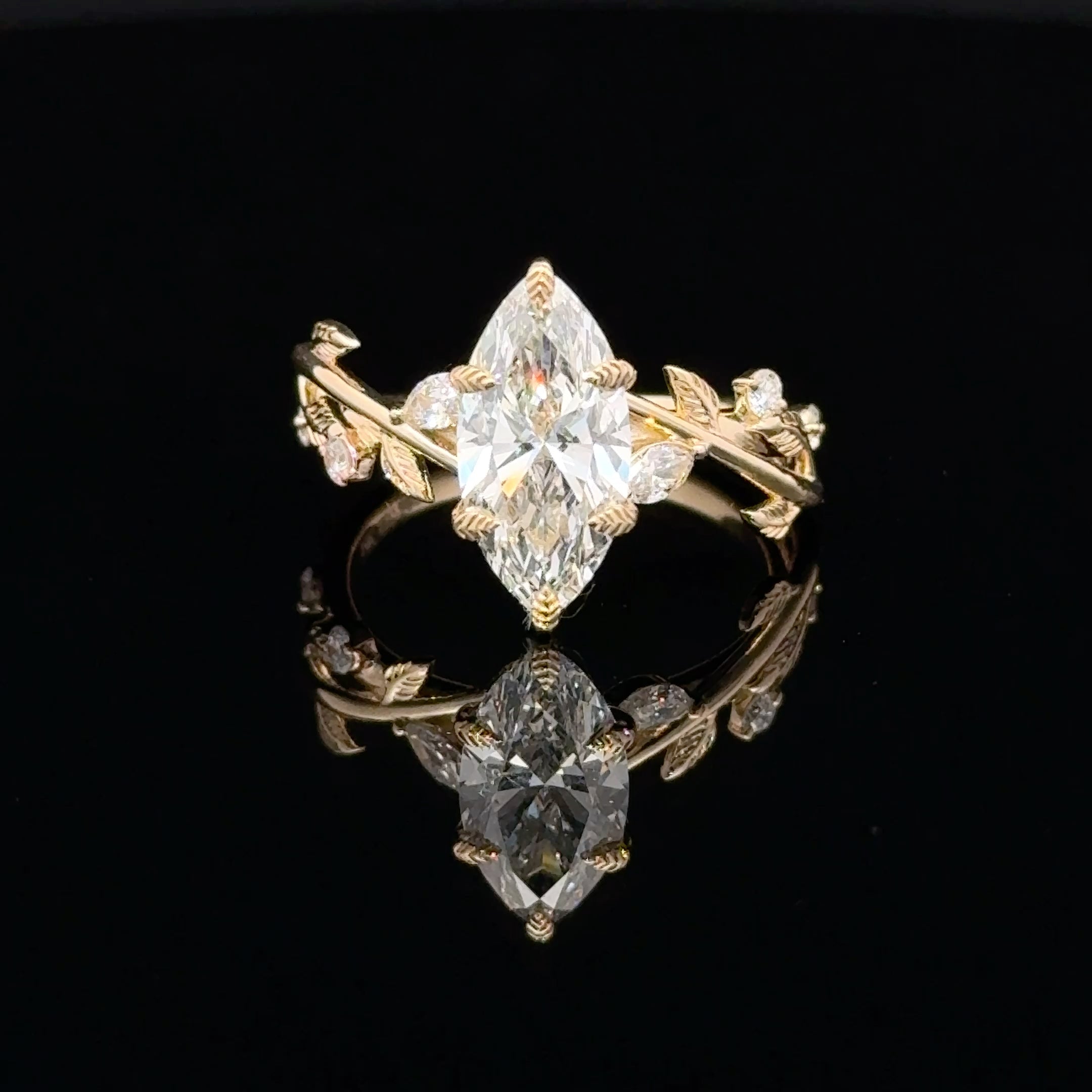 A gold ring featuring a large marquise-cut clear gemstone in the center, with smaller side stones and leaf-like details on a black reflective background.