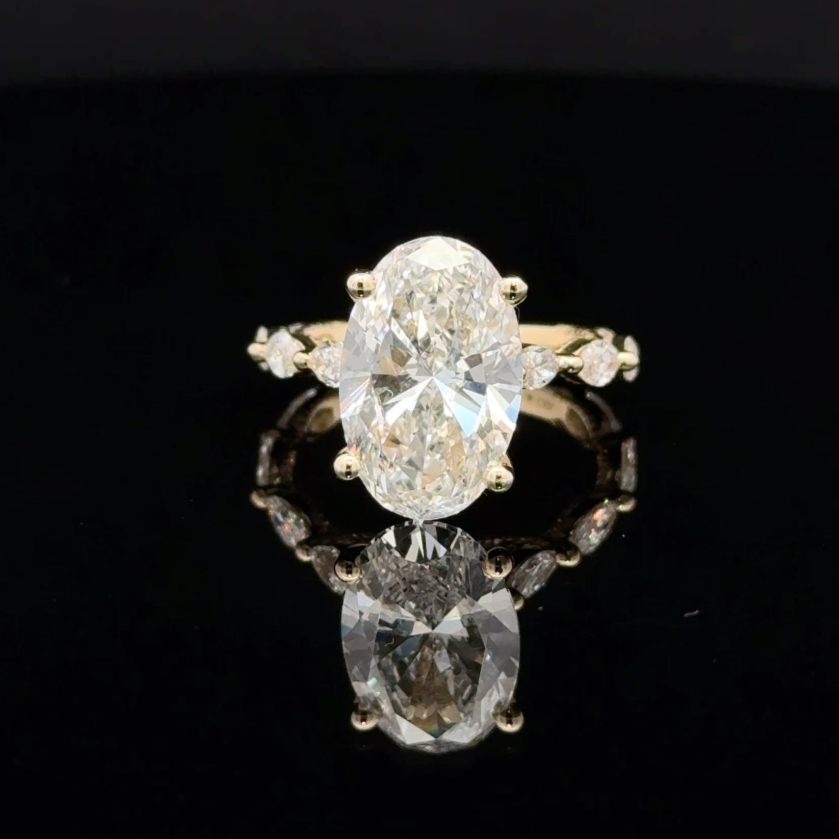 A yellow gold ring featuring a large oval-cut clear center stone with smaller side stones, displayed on a reflective black surface.
