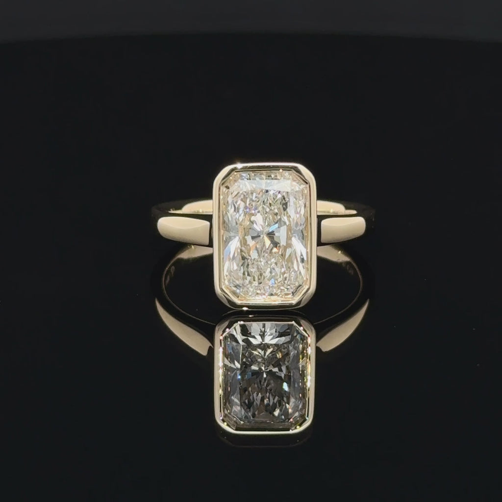 Front view of a 3 carat rectangular radiant-cut diamond in a smooth yellow-gold-toned bezel setting with a polished plain band.