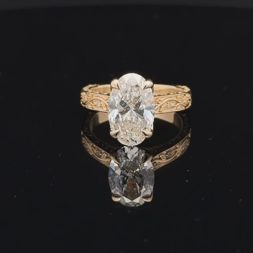 3 carat oval diamond ring with four claw prongs and a gold-toned band featuring winding vines, leaf details and small sparkling accents.