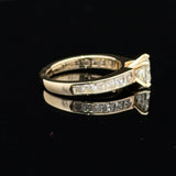 A yellow gold diamond ring with a triangular-cut center stone and channel-set diamonds along the band, displayed on a reflective black surface.