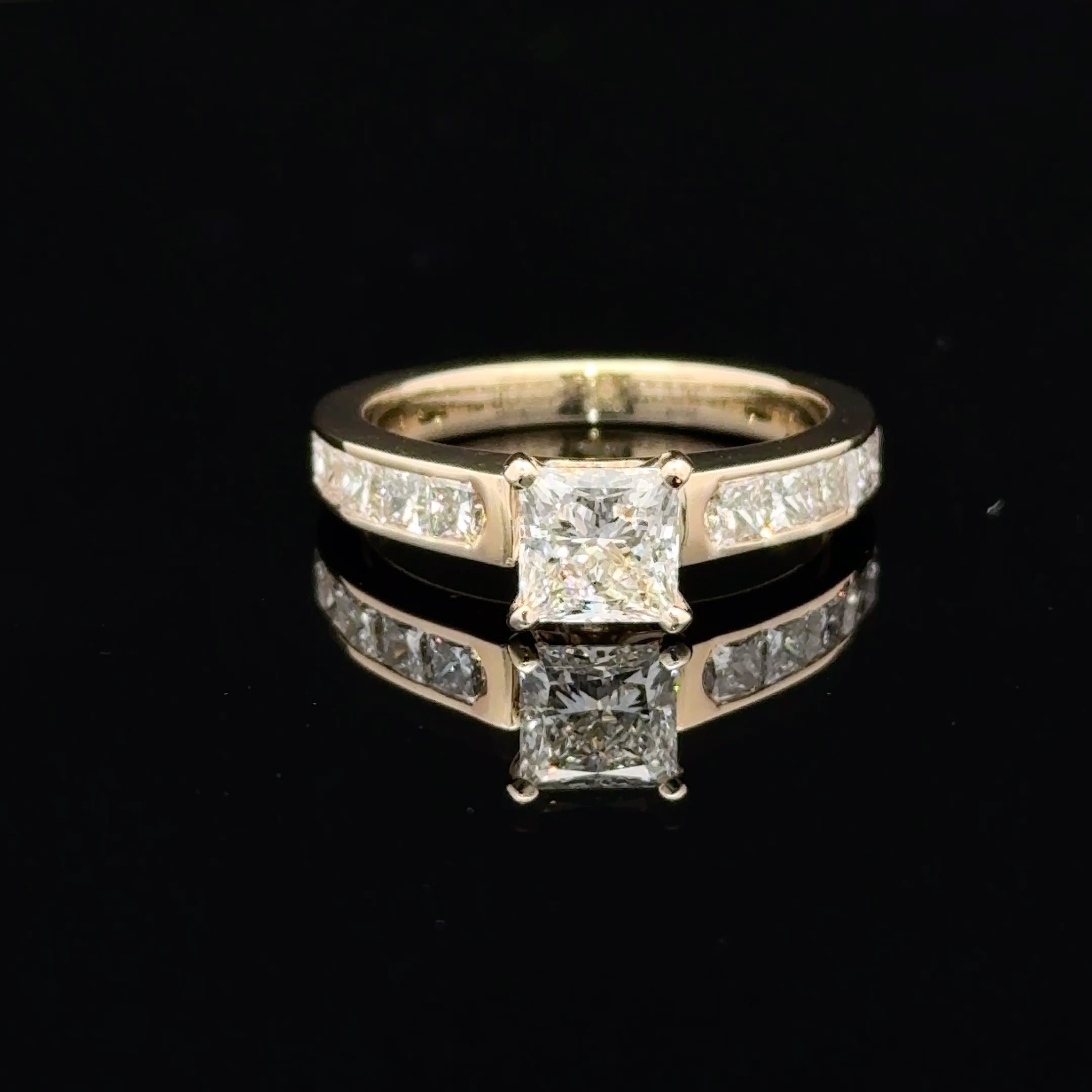 A yellow gold ring featuring a princess-cut diamond center stone and channel-set diamonds along the band, displayed on a reflective black surface.
