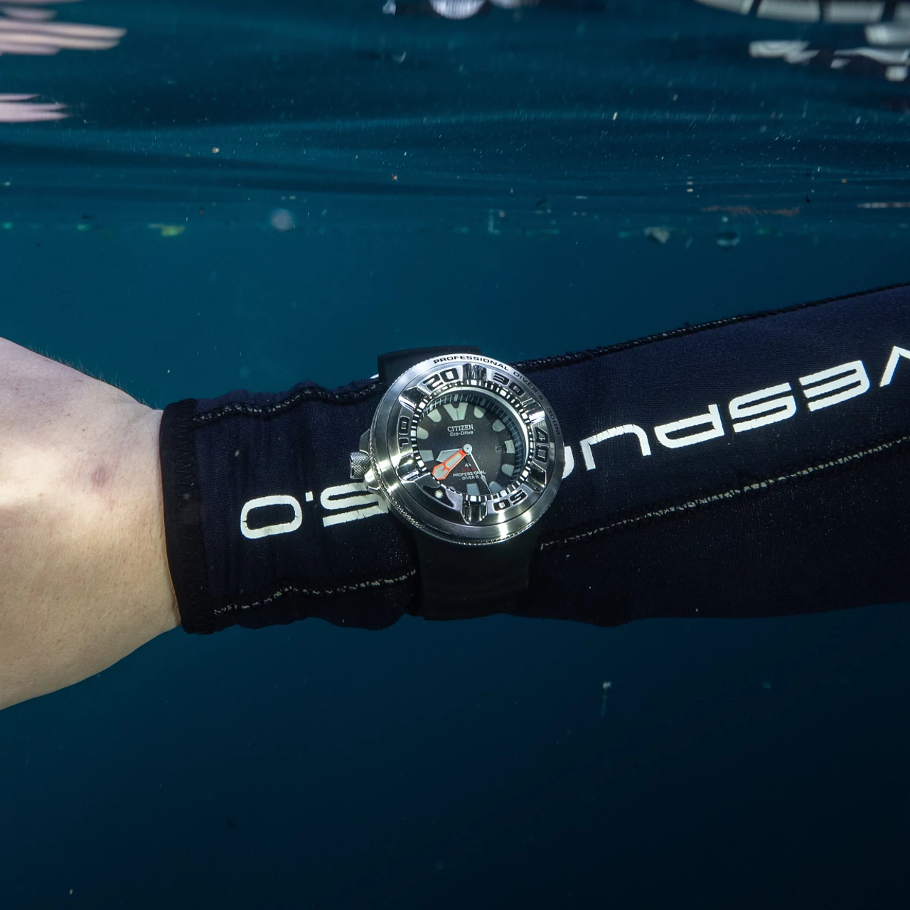 A wristwatch with a black strap and silver-toned case worn on a person's wrist, with a black neoprene wristband featuring white Vespuco branding. The scene is underwater, with blue water and reflections visible.