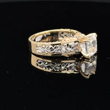 A yellow gold ring with ornate filigree detailing and a clear marquise-cut gemstone, displayed on a reflective black surface.